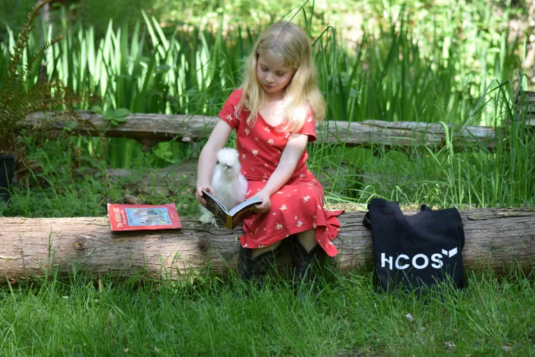 a young girl sitting on a log reading a book to a chicken